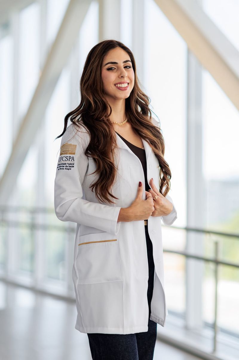 Dra. Camila Medeiros, biomédica, sorrindo, de jaleco branco da UFCSPA, num corredor iluminado.